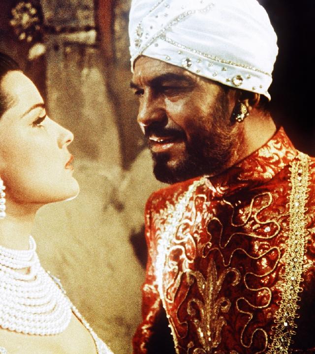 Ein dunkelhäutiger, bärtiger Mann mit weißem Turban nähert sich mit spöttischem Blick von rechts einer festlich hell gekleideten Frau mit halblangem braunem Haar, die mit zahlreichen prunkvollen Perlenketten geschmückt ist und ihn eingeschüchtert anblickt.