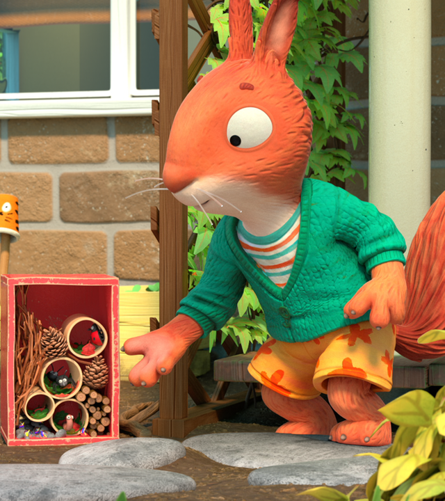 Draußen vor dem Haus: Jamila, das Eichhörnchen, steht rechts neben einem von ihr selbstgebauten Insektenhotel, das aus einer rechteckigen, roten Kiste besteht und mit Stöcken, Zapfen und leeren Rollen gefüllt ist und schaut sich dieses mit stolzem Blick an. Das Kaninchen Pip steht links davon mit gebeugten Knien auf denen er seine Hände hat und schaut mit erstauntem Blick zu Jamila. Im Insektenhotel befinden sich bereits einige Insekten.