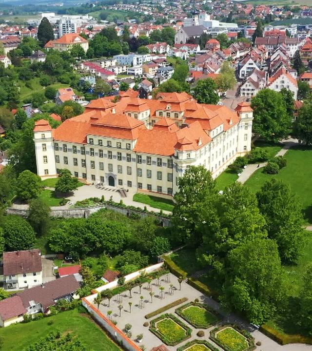 Das Bild zeigt das Schloss Tettnang in einer anschaulichen Perspektive. Das Schloss hat eine helle Fassade und ein markantes, rot gedecktes Dach mit mehreren Erkern und Türmen. Vor dem Schloss erstreckt sich ein gepflegter Garten mit symmetrischen Blumenbeeten, während um das Schloss herum Rasenflächen und Bäume zu sehen sind.  Im Hintergrund sind Teile der Stadt Tettnang zu erkennen, mit vielen kleinen Gebäuden und Wohnhäusern, die ebenfalls rote Dächer besitzen. Diese Gebäude sind in unterschiedlicher Höhe angeordnet und einige sind von Bäumen umgeben. Von dieser Perspektive aus bietet sich auch ein Blick auf die hügelige Landschaft und den klaren Himmel im Hintergrund.  Das Bild vermittelt ein Gefühl von historischer Pracht und zeigt die Verbindung zwischen der Natur und der Architektur des Barockzeitalters.