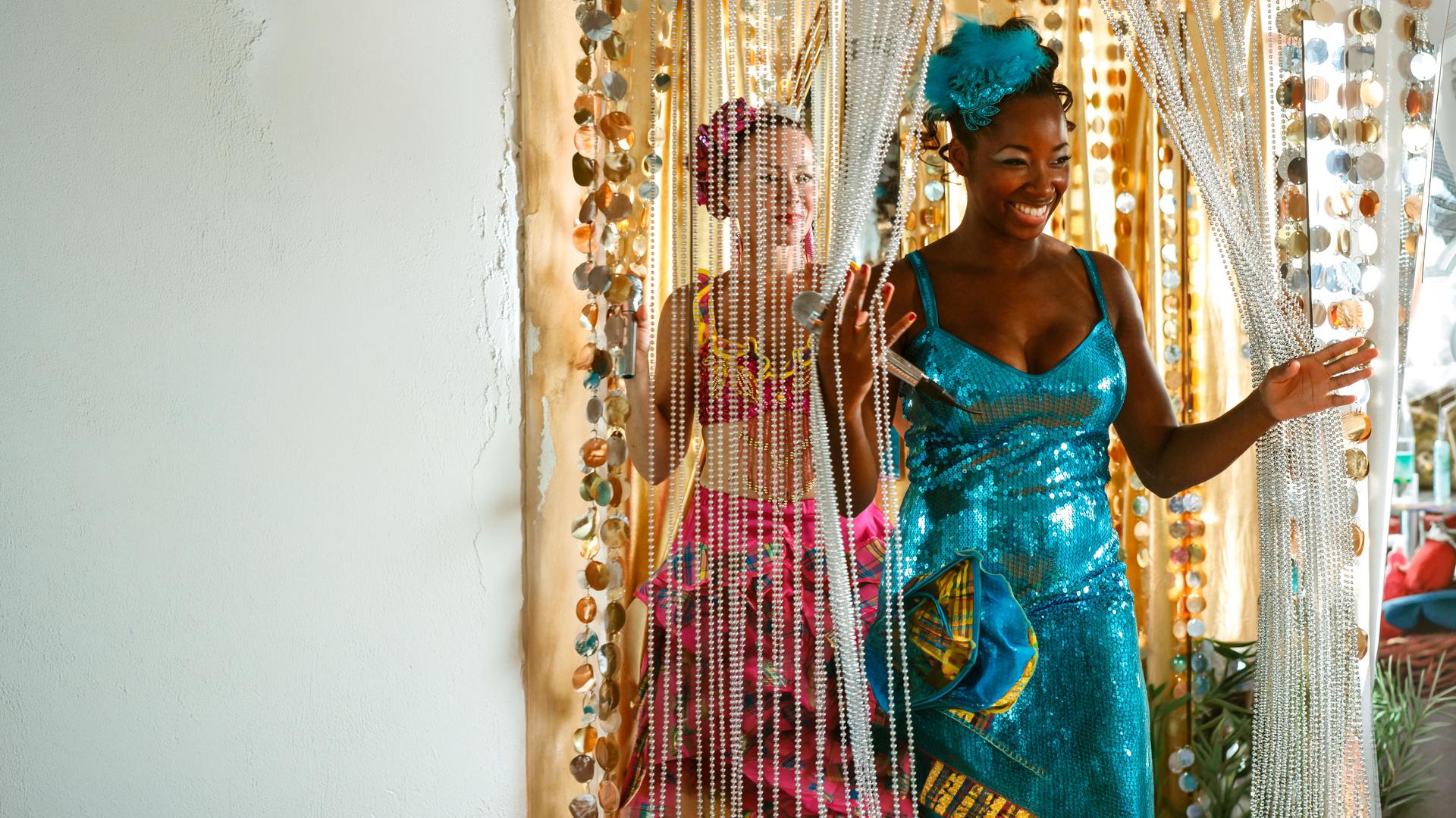 "Death in Paradise - Das letzte Lied": Aimee (Jamelia) geht lächelnd durch einen Perlenvorhang hindurch, sie trägt ein Paillettenkleid, Kopfschmuck und hält ein Mikrofon in der Hand. Hinter ihr steht Eloise (Amanda Mealing), die ähnlich gekleidet ist und in Aimees Richtung schaut.