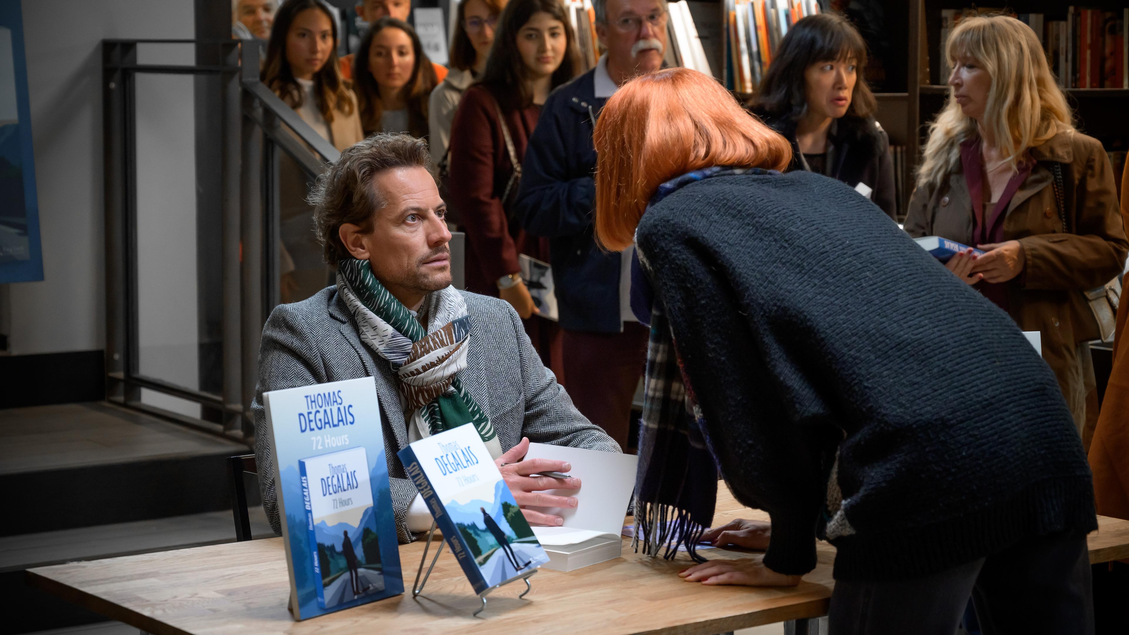 "Das Mädchen und die Nacht (1)": Autor Thomas (Ioan Gruffudd) sitzt bei seiner Autogrammstunde mit verblüfftem Gesichtsausdruck an einem Tisch. Eine junge Frau mit rothaariger Perücke beugt sich zu ihm vor. Im Hintergrund stehen viele Menschen in einer Schlange um sich ein Autogramm zu holen.