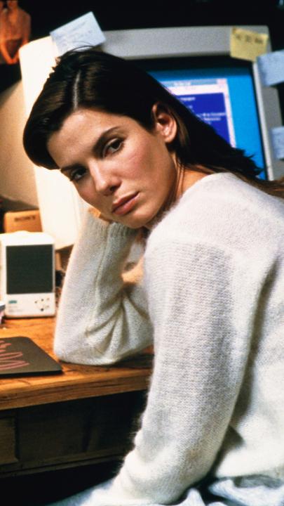 "Das Netz": Die Softwareentwicklerin Angela Bennett (Sandra Bullock) sitzt in ihrem Büro zu Hause, vor sich zwei mit Haftnotizen übersäte Computer, und schaut nachdenklich zur Seite.