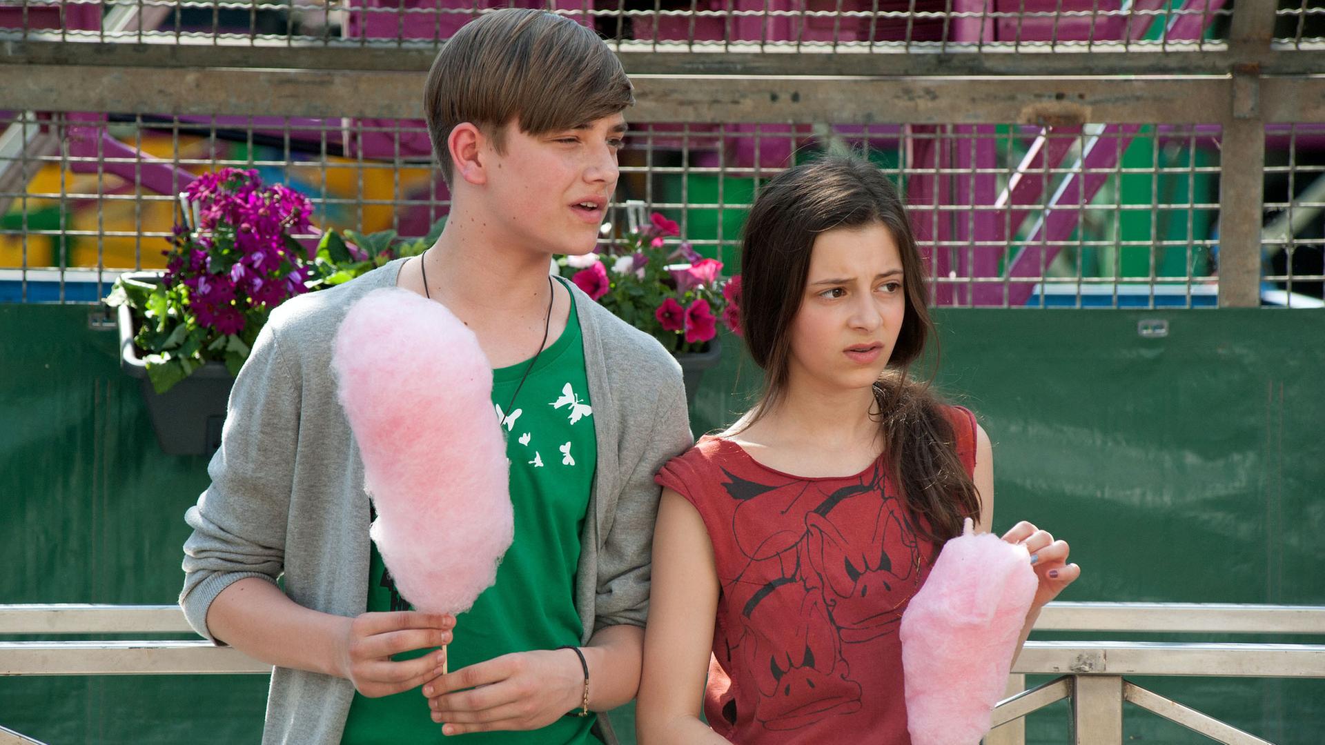 "Das Pubertier - Die Serie: Das Pubertier geht fremd": Lukas (Marc Sierzputowski) steht neben Carla (Mia Kasalo) auf dem Rummel. Beide haben rosafarbene Zuckerwatte in der Hand.