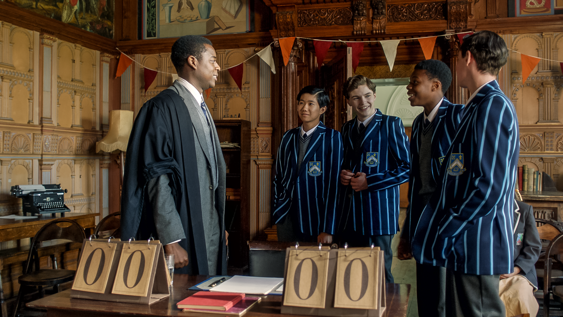 Mr. Parker (links) steht neben den vier Jungs Clifford (2. von links), Sebastian (Mitte), Victor (2. von rechts) und Cyril (rechts) vom Thackerton College und lächelt diese an. Die Jungs grinsen mit süffisantem Lächeln zurück.  Im Raum hängen rote und weiße Wimpel. Auf einem Pult stehen zwei Punktetafeln für das bevorstehende Quiz. Noch lautet der Punktestand Null zu Null.