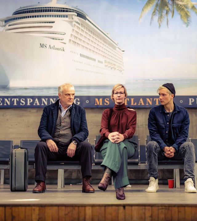 Auf einem Bahnsteig sitzen die Protagonisten Nils und Vincent wartend auf Stühlen, sie sehen eine Frau an, die zwischen ihnen sitzt. Hinter ihnen an der Wand hängt eine Reklame für eine Kreuzfahrt.