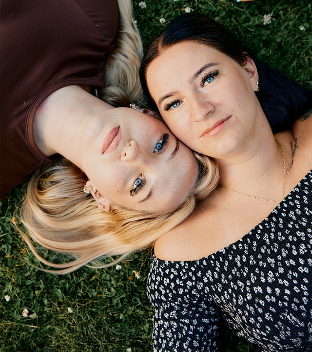 Blick von oben: Jaqueline und Caithlen liegen auf dem Rasen und berühren sich an der Hand.
