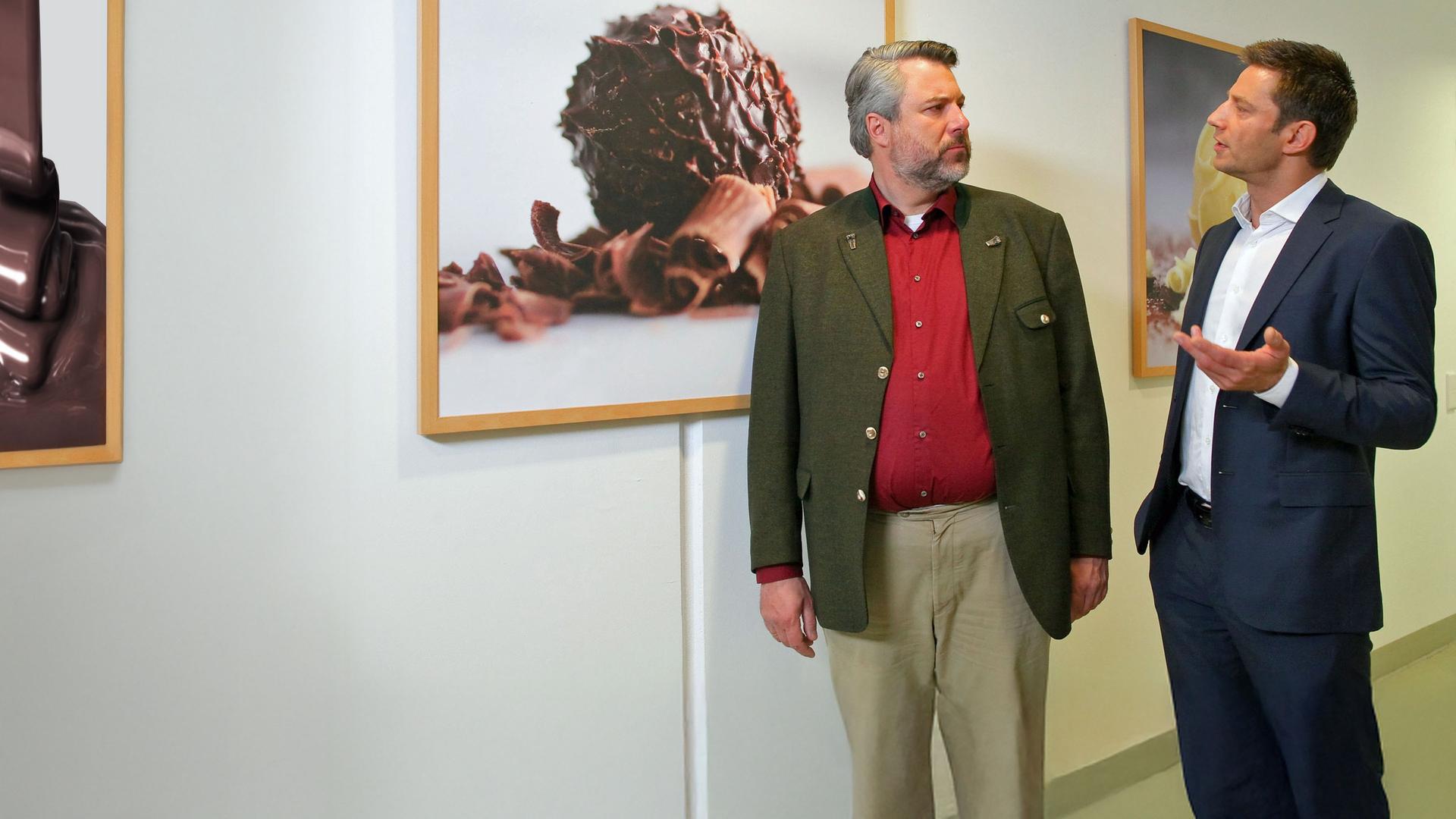 "Die Rosenheim-Cops - Das süße Erbe": Die Kommissare Stadler (Dieter Fischer) und Hansen (Igor Jeftic) unterhalten sich auf einem Flur. Hinter ihnen an der Wand sind gerahmte Fotoleinwände mit Trüffelpralinen abgebildet.
