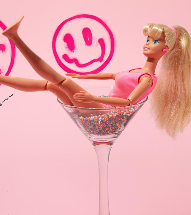 Eine Barbiepuppe liegt in einem Cocktailglas. Der rosane Hintergrund hat Risse und aufgesprühte Graffiti-Smileys