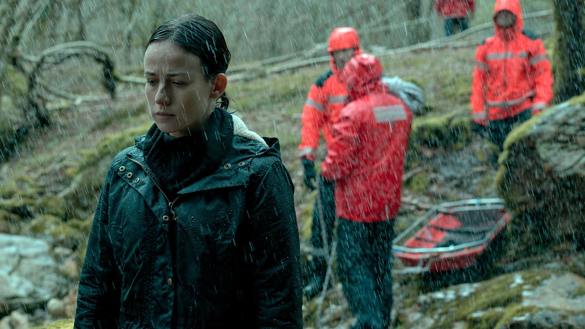 Amaia Salazar (Marta Etura) steht mit geschlossenen Augen im Wald. Es regnet und im Hintergrund sind rot gekleidete Rettungskräfte zu sehen.