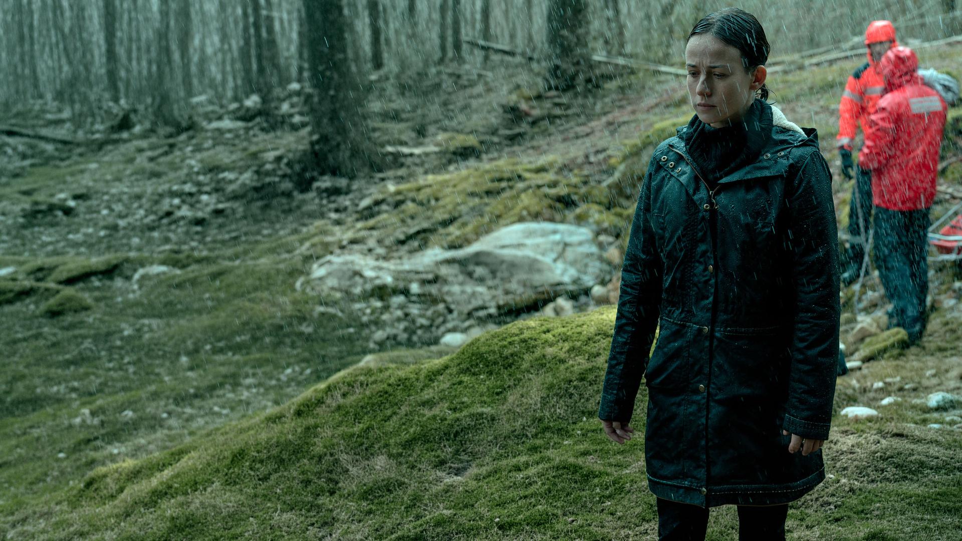 Amaia Salazar (Marta Etura) steht mit geschlossenen Augen im Wald. Es regnet und im Hintergrund sind rot gekleidete Rettungskräfte zu sehen.
