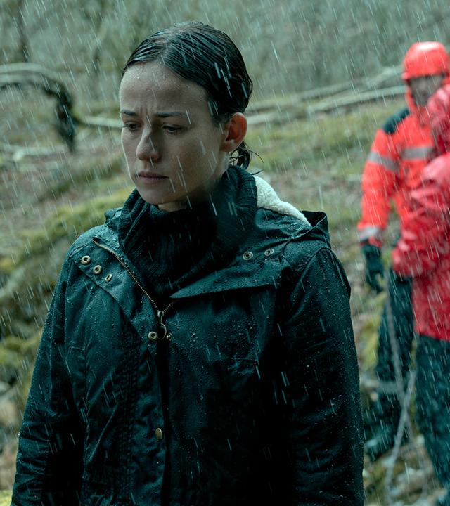Amaia Salazar (Marta Etura) steht mit geschlossenen Augen im Wald. Es regnet und im Hintergrund sind rot gekleidete Rettungskräfte zu sehen.