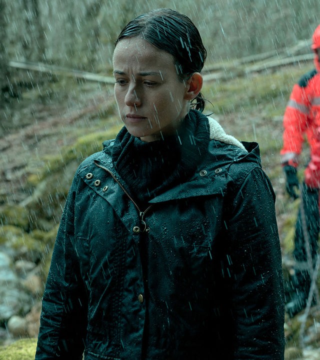 Amaia Salazar (Marta Etura) steht mit geschlossenen Augen im Wald. Es regnet und im Hintergrund sind rot gekleidete Rettungskräfte zu sehen.