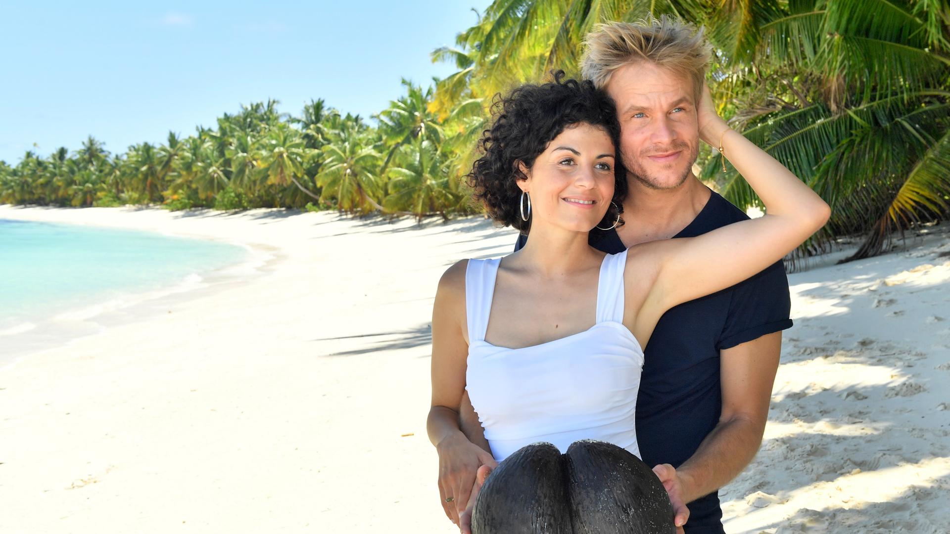 Sebastian Schuster (Frederic Heidorn) und Ariane Hoffmann (Liza Tzschirner) stehen am Sandstrand. Sebastian umarmt Ariane von hinten und hält dabei eine Kokosnuss in den Händen. Sie streicht ihm dabei durchs Haar.
