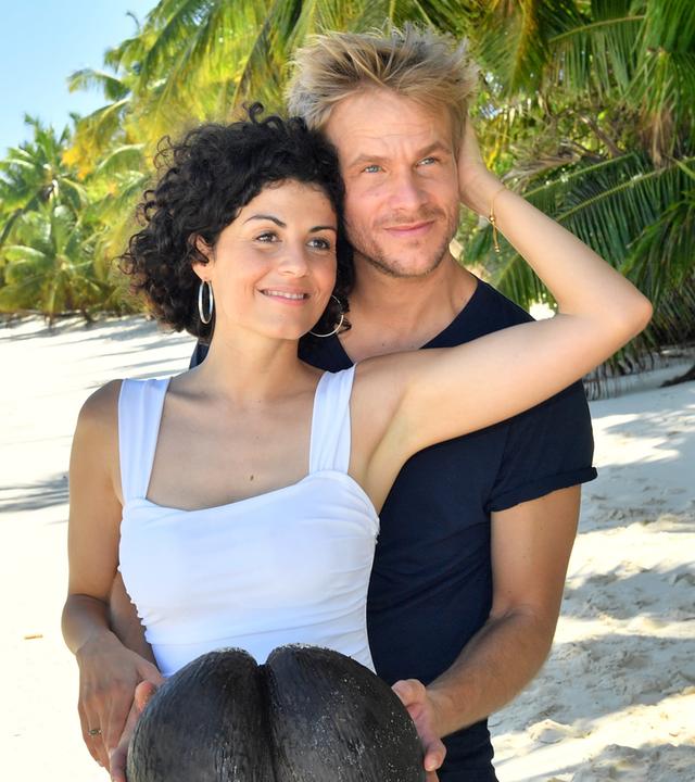 Sebastian Schuster (Frederic Heidorn) und Ariane Hoffmann (Liza Tzschirner) stehen am Sandstrand. Sebastian umarmt Ariane von hinten und hält dabei eine Kokosnuss in den Händen. Sie streicht ihm dabei durchs Haar.