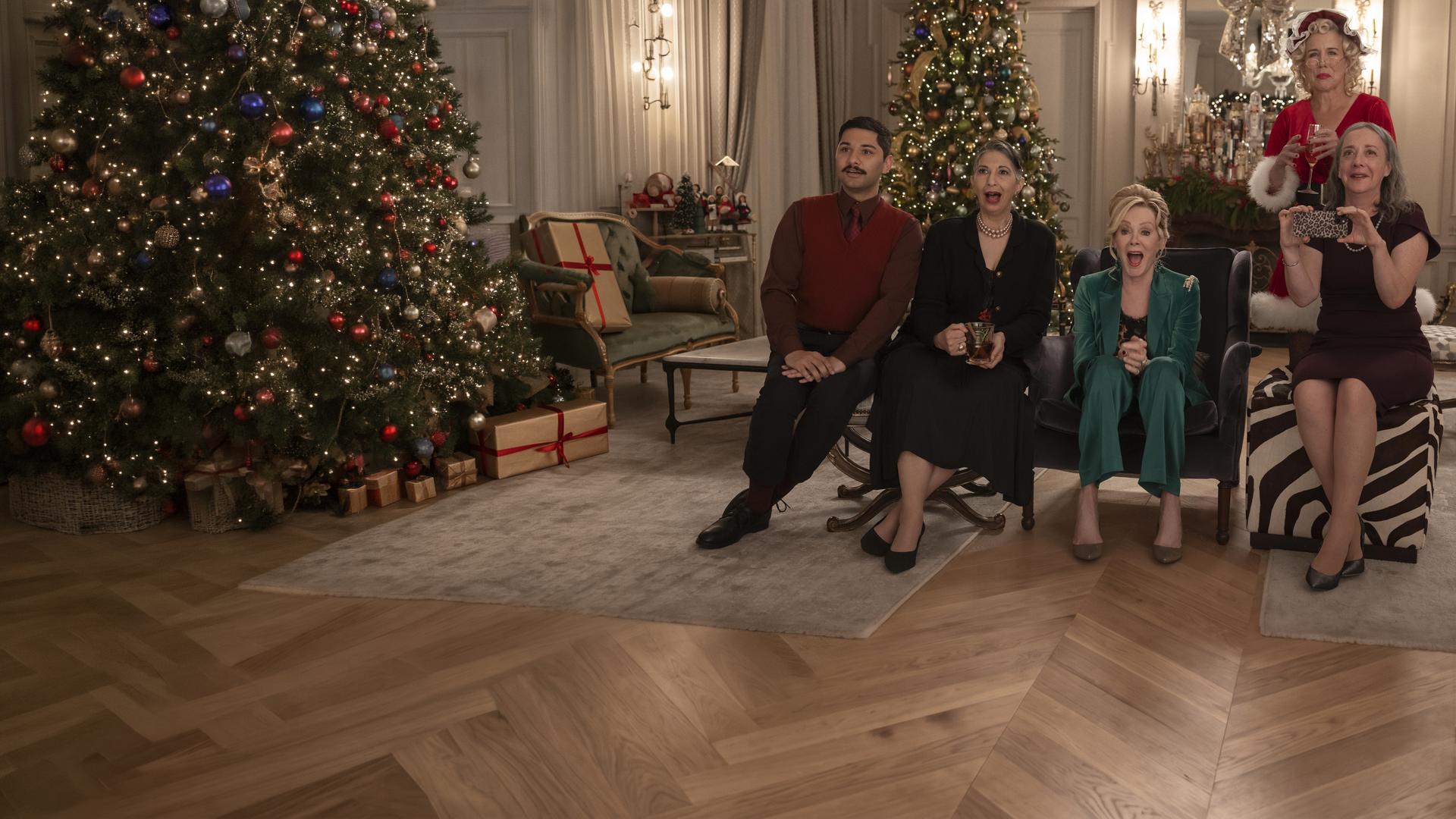 "Hacks - Das Vegas-Weihnachtsspektakel": In festlicher Kleidung sitzen Damien (Mark Indelicato), Josefina (Rose Abdoo), Deborah (Jean Smart) und Avas Mutter Nina (Jane Adams) lachend nebeneinander in einem festlich geschmückten Wohnraum. Hinter ihnen steht Bürgermeisterin Mayor Jo (Lauren Weedman) im Santa-Kostüm. An der Seite steht ein großer Weihnachtsbaum.