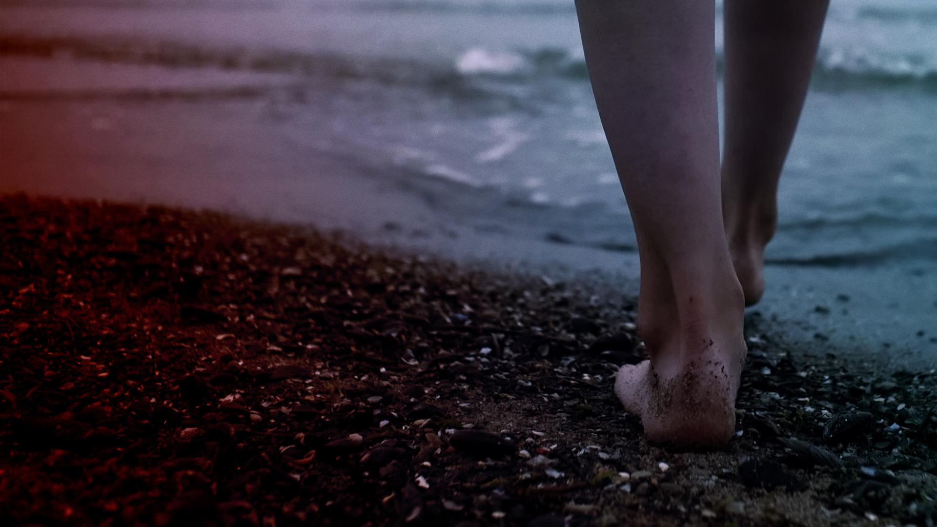 Zwei nackte Beine gehen über einen Kiesstrand auf das Wasser zu.