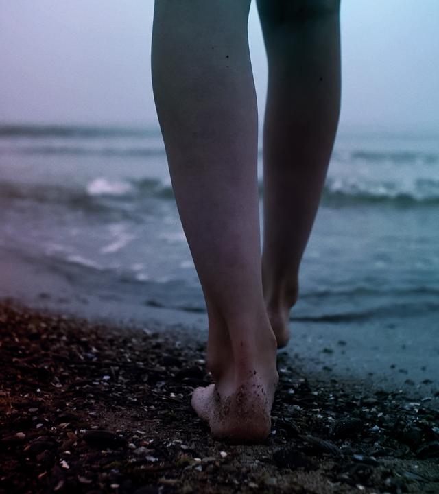 Zwei nackte Beine gehen über einen Kiesstrand auf das Wasser zu.
