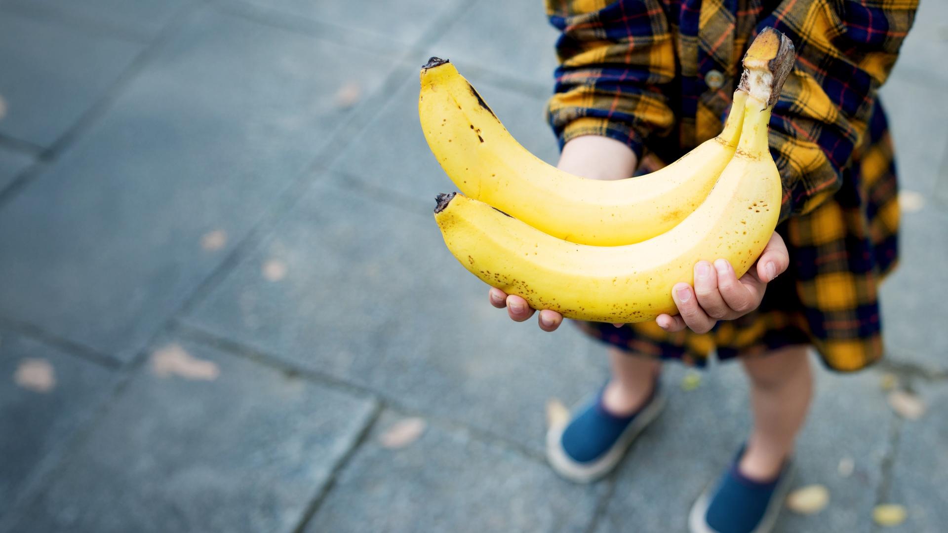 Blick von schräg oben auf zwei gelbe Bananen in den Händen eines Kindes.
