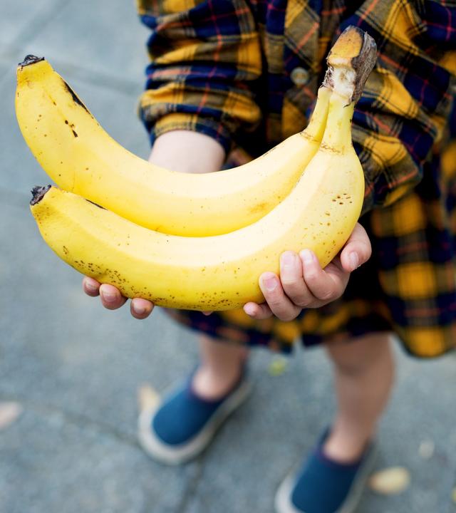 Blick von schräg oben auf zwei gelbe Bananen in den Händen eines Kindes.