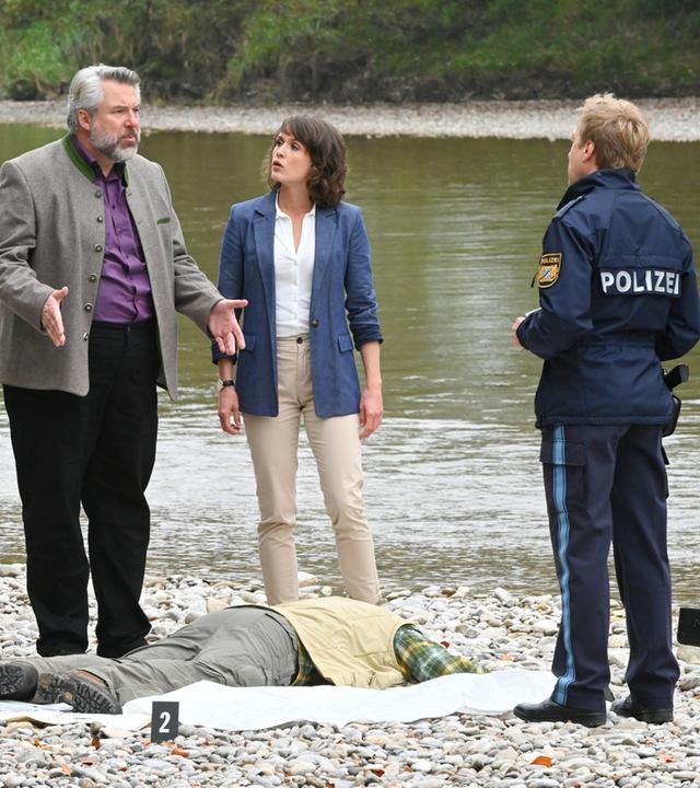 "Die Rosenheim-Cops - Das Wasser zum Glück": Das Ermittlerteam der Kripo Rosenheim, Pathologin Atay (Sevda Polat), Kommissar Stadler (Dieter Fischer), Kommissarin Andresen (Sophie Melbinger) und Polizeihauptmeister Mohr (Max Müller), steht am Ufer eines Flusses, vor ihnen liegt das Opfer (Komparse) bäuchlings im Kies.