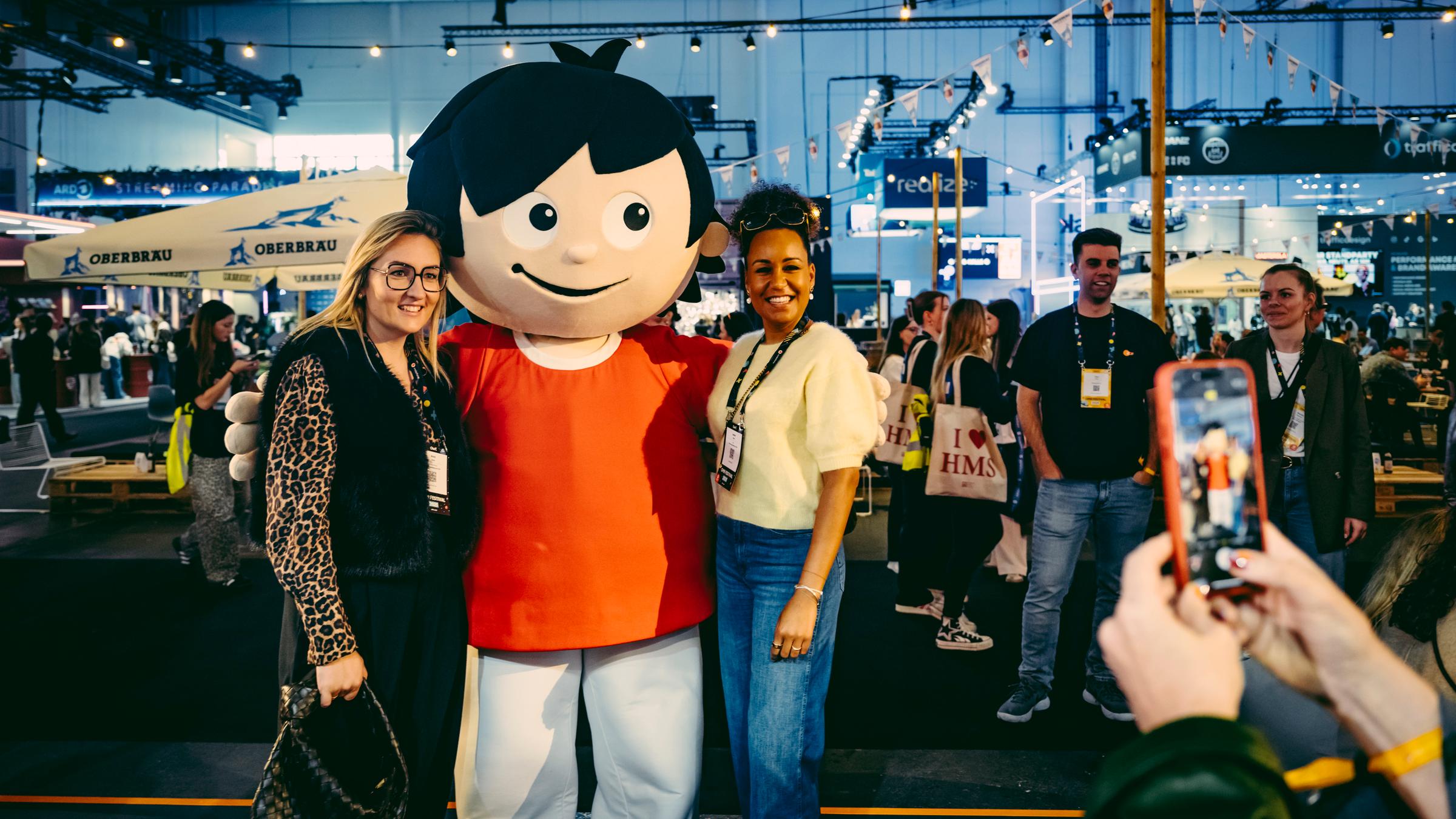Zwei Messebesucherinnen der OMR 2025 in Hamburg stellen sich mit einem Mainzelmännchen zum gemeinsamen beneinander zum Foto.