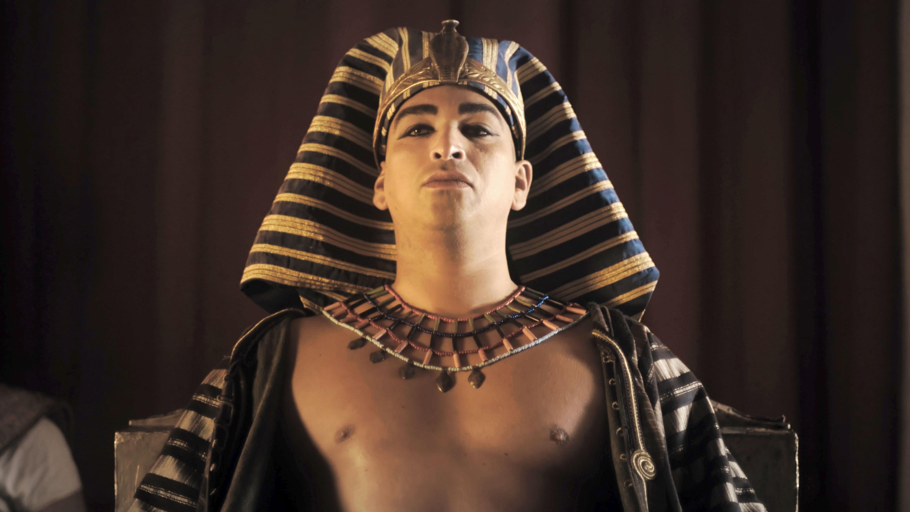  Spielszene. Pharao Snofru, auf dem Kopf ein gold-blau quergestreiftes Nemes-Kopftuch mit goldenem Stirnring mit einer Kobra. Um den Hals ein breiter mit bunten Steinen verzierter Goldkragen. Die Brust ist nackt, über den Schultern hängt ein gold-blau quergestreifter Umhang.