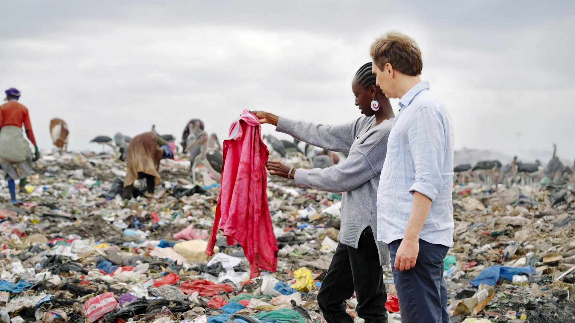 Ein Mann und eine Frau stehen in einer Mülldeponie zwischen Menschen und Tieren, die im Müll suchen.