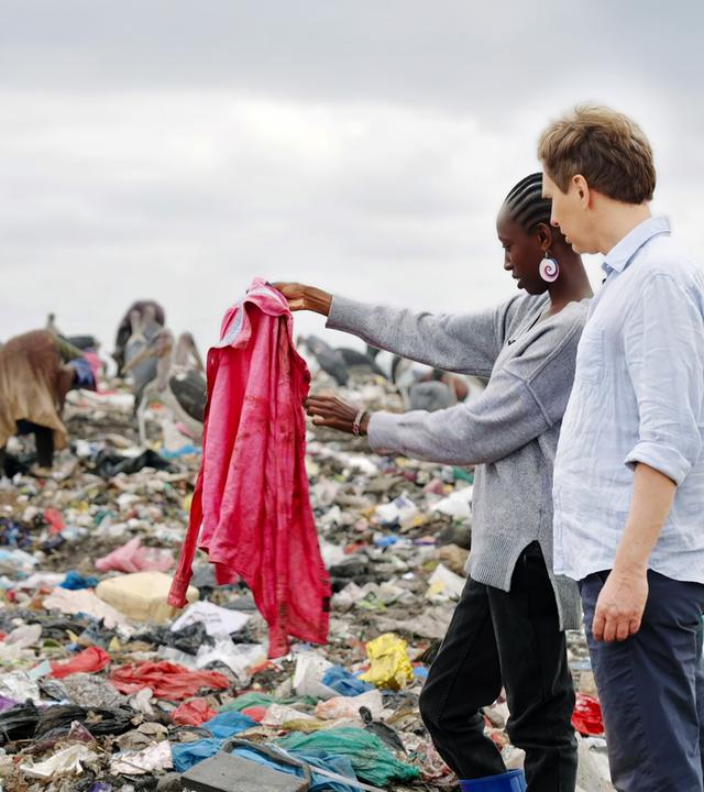 Ein Mann und eine Frau stehen in einer Mülldeponie zwischen Menschen und Tieren, die im Müll suchen.