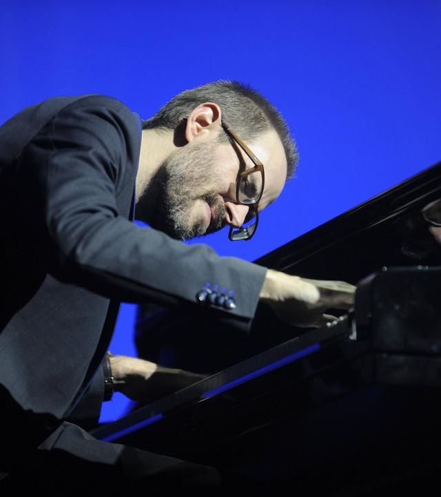 Ein Mann mit Brille und Bart beugt sich über ein Klavier und spielt.