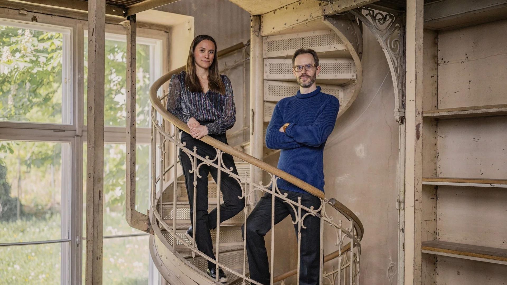 Zwei Musiker stehen in einem Innenraum mit Instrumenten; das Duo ist nebeneinander positioniert, die Umgebung wirkt wie ein Studio oder Proberaum mit sichtbaren Details im Hintergrund.