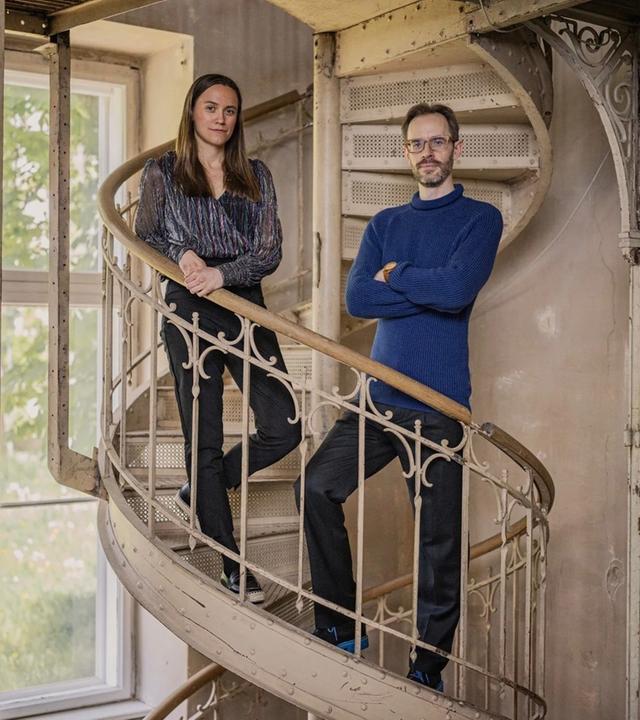 Zwei Musiker stehen in einem Innenraum mit Instrumenten; das Duo ist nebeneinander positioniert, die Umgebung wirkt wie ein Studio oder Proberaum mit sichtbaren Details im Hintergrund.