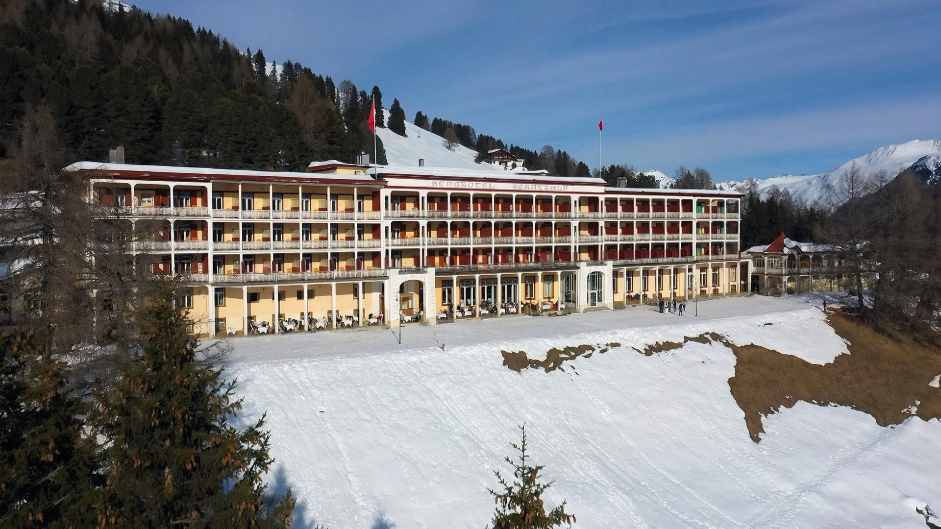 Das Bild zeigt das Bergrestaurant Schatzalp in Davos, Schweiz. Es handelt sich um ein mehrstöckiges Gebäude aus der Zeit um 1900, das in einem hellen Gelb und Weiß gehalten ist. Die Fassade ist von mehreren Balkonen mit schmiedeeisernen Geländern geprägt. Im Erdgeschoss sind große Fenster zu sehen, vor denen Tischchen stehen, auf denen Stühle platziert sind. An der Vorderseite des Gebäudes gibt es einen Eingangsbereich, der durch einen großen Bogen gekennzeichnet ist.  Um das Hotel herum liegt eine schneebedeckte Landschaft mit Nadelbäumen im Vordergrund. Im Hintergrund erheben sich schneebedeckte Berge unter einem leicht bewölkten Himmel. Einige Personen sind auf dem Platz vor dem Hotel zu sehen, während andere im Hintergrund zwischen den Bäumen stehen. Das gesamte Umfeld vermittelt eine ruhige, winterliche Atmosphäre.