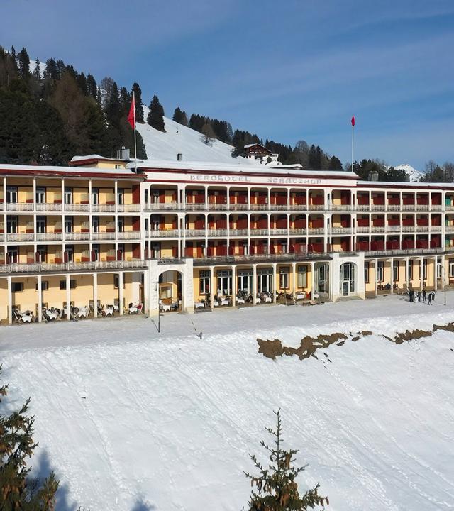 Das Bild zeigt das Bergrestaurant Schatzalp in Davos, Schweiz. Es handelt sich um ein mehrstöckiges Gebäude aus der Zeit um 1900, das in einem hellen Gelb und Weiß gehalten ist. Die Fassade ist von mehreren Balkonen mit schmiedeeisernen Geländern geprägt. Im Erdgeschoss sind große Fenster zu sehen, vor denen Tischchen stehen, auf denen Stühle platziert sind. An der Vorderseite des Gebäudes gibt es einen Eingangsbereich, der durch einen großen Bogen gekennzeichnet ist.  Um das Hotel herum liegt eine schneebedeckte Landschaft mit Nadelbäumen im Vordergrund. Im Hintergrund erheben sich schneebedeckte Berge unter einem leicht bewölkten Himmel. Einige Personen sind auf dem Platz vor dem Hotel zu sehen, während andere im Hintergrund zwischen den Bäumen stehen. Das gesamte Umfeld vermittelt eine ruhige, winterliche Atmosphäre.