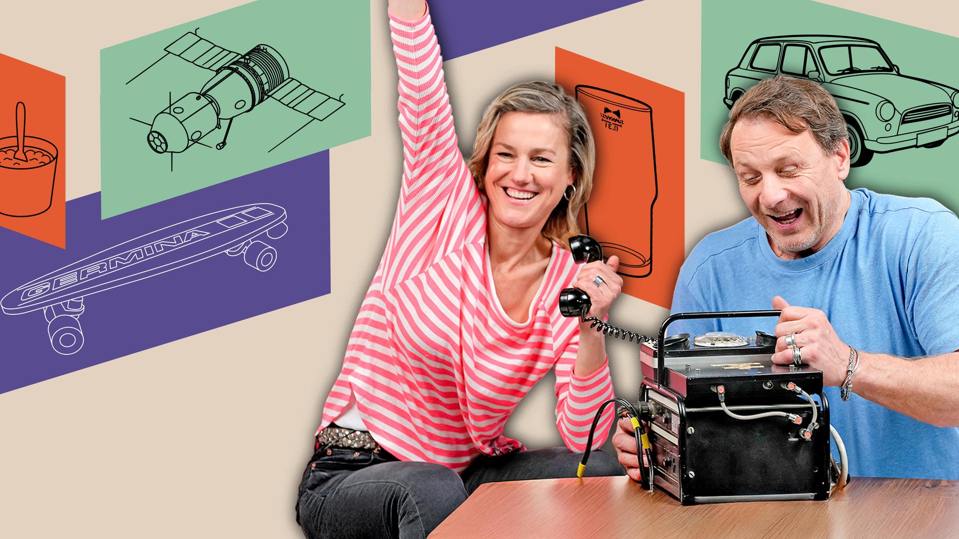 Rhea Harder-Vennewald und Hendrik Duryn sitzen lachend auf roten Sesseln vor der schwarzen Box eines Funktelefons, das auf einem kleinen Tisch platziert ist. Er hantiert mit der Box, während sie den Hörer in der Hand hält und den anderen Arm triumphierend in die Höhe reckt.