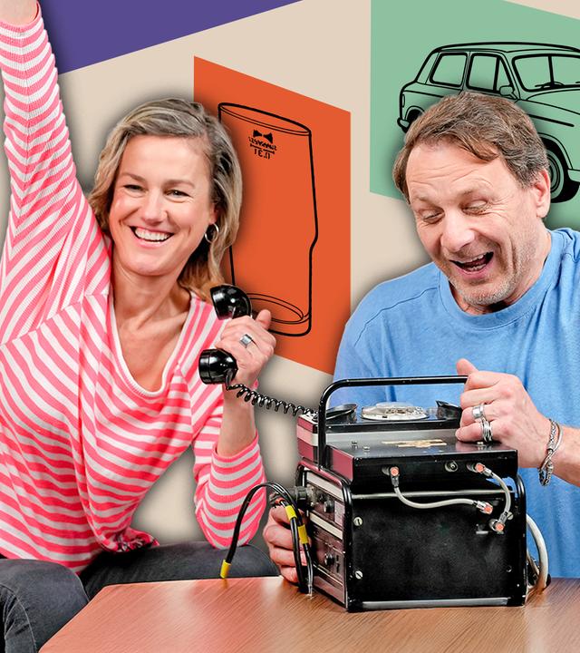 Rhea Harder-Vennewald und Hendrik Duryn sitzen lachend auf roten Sesseln vor der schwarzen Box eines Funktelefons, das auf einem kleinen Tisch platziert ist. Er hantiert mit der Box, während sie den Hörer in der Hand hält und den anderen Arm triumphierend in die Höhe reckt.