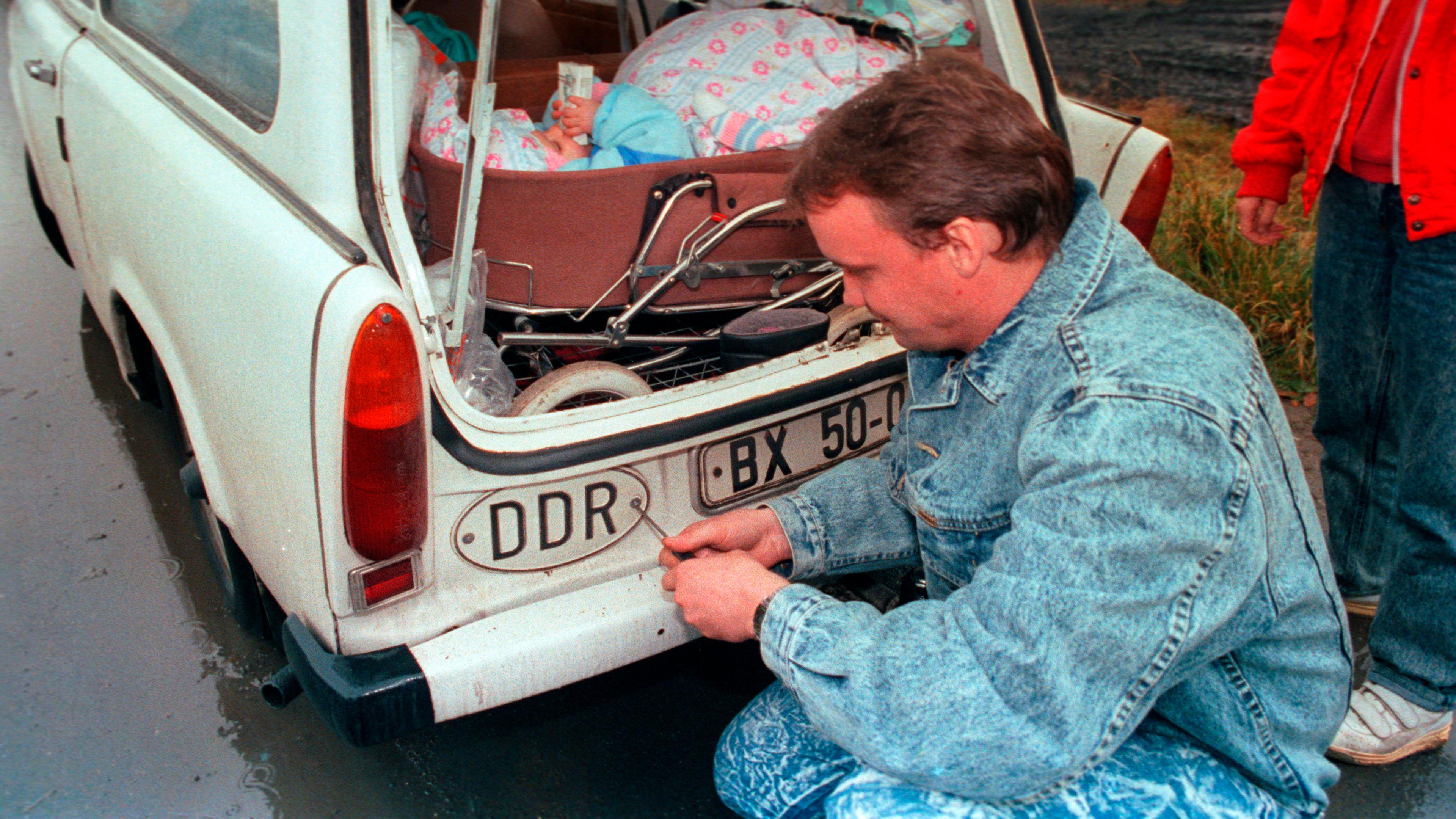 Ein Mann entfernt mit einem Schraubendreher das Länderkennzeichen DDR von seinem Trabant.