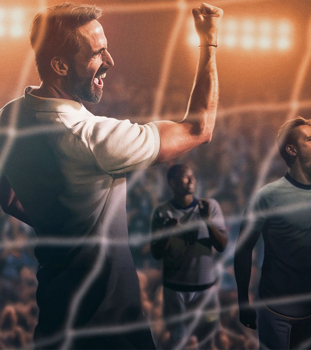 "Dear England (1)": Stilisiertes Bild, im Stadion: Gareth Southgate (Joseph Fiennes) trägt ein weißes Poloshirt und hat den den rechten, muskulösen Arm mit geballter Faust jubelnd in die Höhe gereckt. Hinter ihm befinden sich einige Spieler, dahinter sitzt Publikum auf den Rängen eines Stadions.