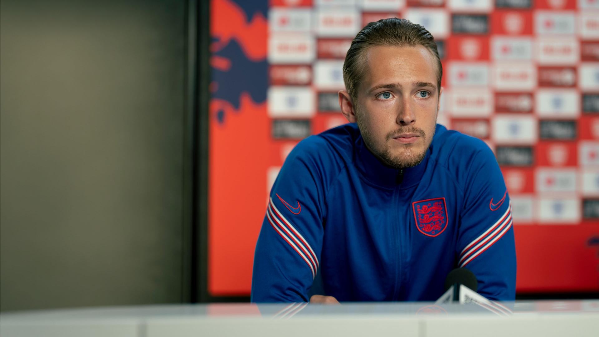 "Dear England (2)": Harry Kane (Will Antenbring) sitzt mit nachdenklichem Gesichtsausdruck an einem Tisch. Er trägt ein überwiegend blaues Trikot mit weiß-roten Streifen auf den Ärmeln und mit England-Emblem.