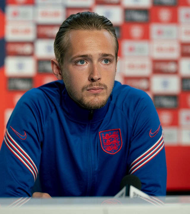 "Dear England (2)": Harry Kane (Will Antenbring) sitzt mit nachdenklichem Gesichtsausdruck an einem Tisch. Er trägt ein überwiegend blaues Trikot mit weiß-roten Streifen auf den Ärmeln und mit England-Emblem.