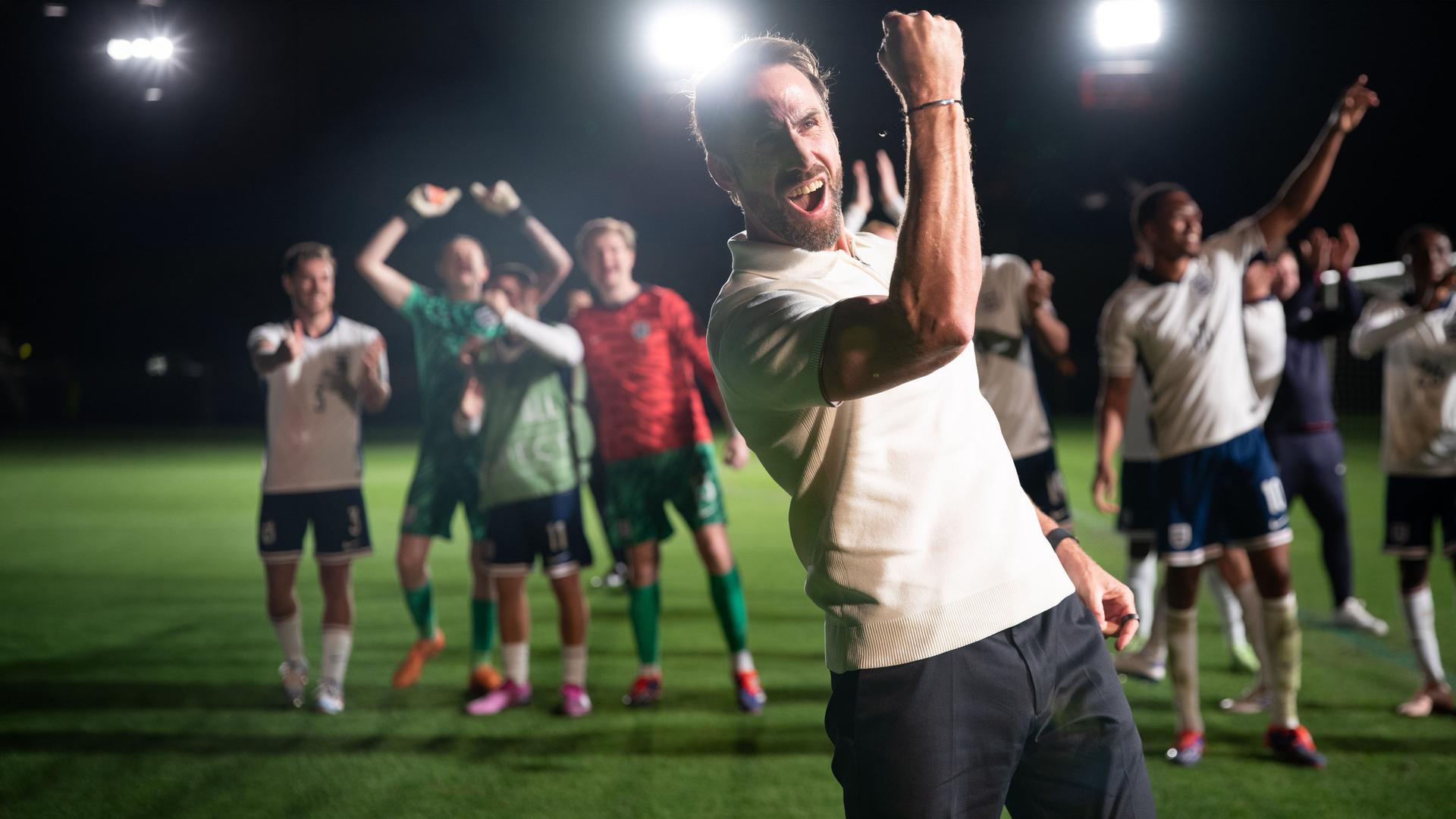 "Dear England (4)": Bei Flutlicht nachts im Stadion auf dem Rasen: Im Vordergrund reckt Gareth Southgate (Joseph Fiennes) seinen rechten Arm mit geballter Faust vor Kraft strotzend in die Höhe. Hinter ihm befindet sich seine Mannschaft, die ebenfalls jubiliert.
