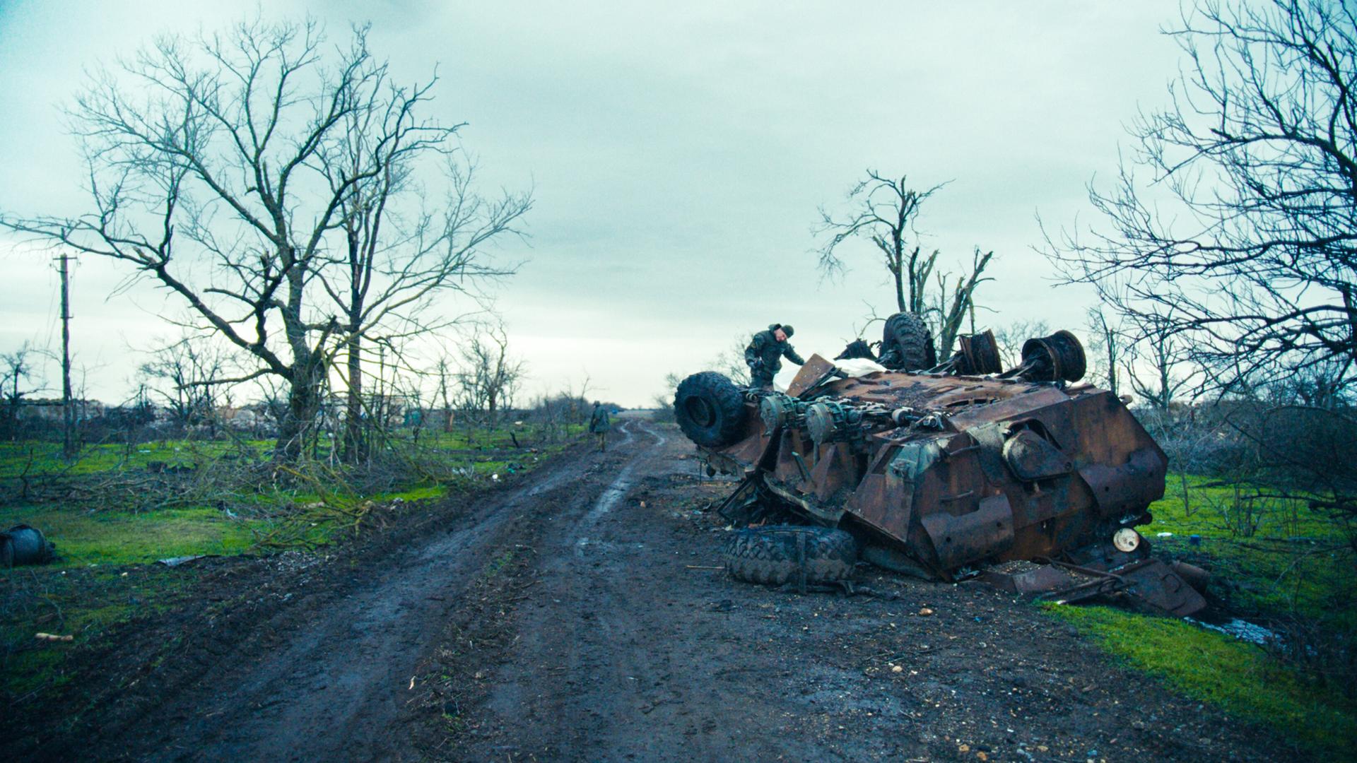 Das Bild zeigt eine trostlose Landschaft, die stark von den Folgen eines Krieges geprägt ist. Im Vordergrund liegt ein umgestürzter Panzer, dessen rostige Oberfläche und beschädigte Teile deutlich sichtbar sind. Auf dem Panzer steht ein Mann, der an dem Fahrzeug arbeitet. Seine Körperhaltung deutet auf eine gewissenhafte Beschäftigung hin. Im Hintergrund sind weitere, kahle Bäume sichtbar, die kaum noch lebendes Geäst tragen. Der Himmel ist bewölkt und erscheint düster, was die traurige Atmosphäre der Szene verstärkt. Die Erde ist matschig und wirkt unordentlich, mit verstreuten Überresten, die auf die Zerstörung in der Umgebung hinweisen. Der Weg, der sich durch die Landschaft schlängelt, ist ebenfalls von Schlamm und Trümmern gezeichnet. Alles in diesem Bild vermittelt ein starkes Gefühl von Verwüstung und den Auswirkungen des Krieges auf das tägliche Leben der Menschen in der Region.