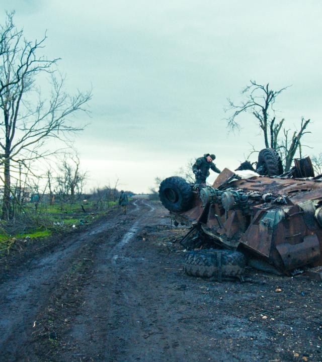 Das Bild zeigt eine trostlose Landschaft, die stark von den Folgen eines Krieges geprägt ist. Im Vordergrund liegt ein umgestürzter Panzer, dessen rostige Oberfläche und beschädigte Teile deutlich sichtbar sind. Auf dem Panzer steht ein Mann, der an dem Fahrzeug arbeitet. Seine Körperhaltung deutet auf eine gewissenhafte Beschäftigung hin. Im Hintergrund sind weitere, kahle Bäume sichtbar, die kaum noch lebendes Geäst tragen. Der Himmel ist bewölkt und erscheint düster, was die traurige Atmosphäre der Szene verstärkt. Die Erde ist matschig und wirkt unordentlich, mit verstreuten Überresten, die auf die Zerstörung in der Umgebung hinweisen. Der Weg, der sich durch die Landschaft schlängelt, ist ebenfalls von Schlamm und Trümmern gezeichnet. Alles in diesem Bild vermittelt ein starkes Gefühl von Verwüstung und den Auswirkungen des Krieges auf das tägliche Leben der Menschen in der Region.