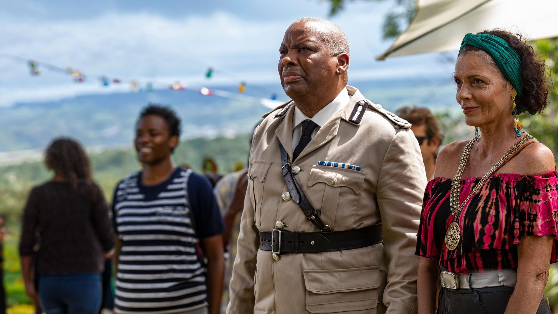 "Death in Paradise - Der Rückkehrer": Commissioner Selwyn Patterson (Don Warrington) und Catherine Bordey (Elizabeth Bourgine) stehen zusammen mit anderen Personen draußen und blicken in Richtung Bühne.