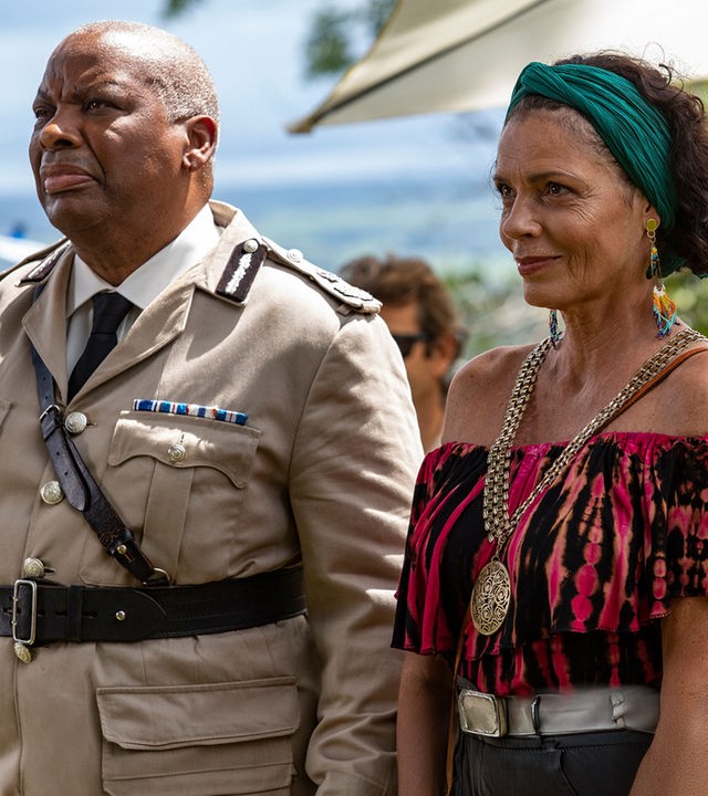 "Death in Paradise - Der Rückkehrer": Commissioner Selwyn Patterson (Don Warrington) und Catherine Bordey (Elizabeth Bourgine) stehen zusammen mit anderen Personen draußen und blicken in Richtung Bühne.