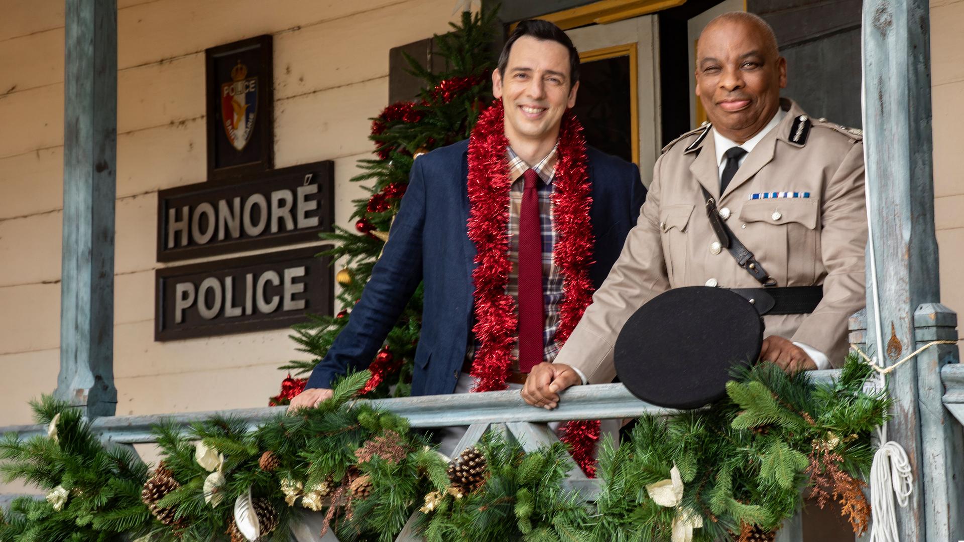 "Death in Paradise - Der Weihnachtsgeist (1)": Detective Neville Parker (Ralf Little) und der Polizeichef Selwyn Patterson (Don Warrington) stehen auf der weihnachtlich dekorierten Veranda der Polizeiwache und schauen zufrieden in die Ferne.