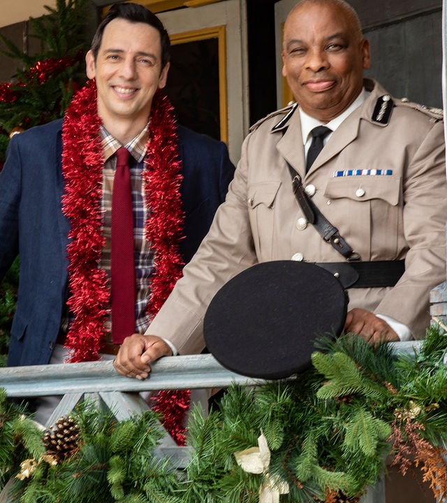 "Death in Paradise - Der Weihnachtsgeist (1)": Detective Neville Parker (Ralf Little) und der Polizeichef Selwyn Patterson (Don Warrington) stehen auf der weihnachtlich dekorierten Veranda der Polizeiwache und schauen zufrieden in die Ferne.