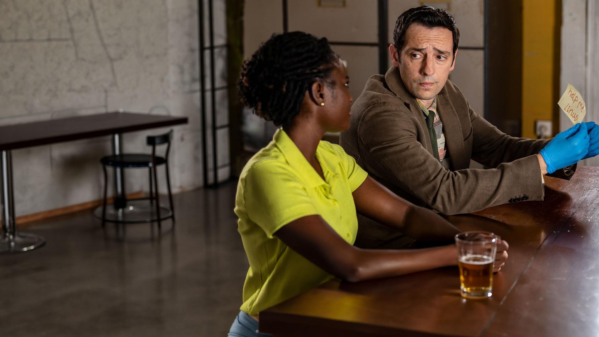 "Death in Paradise - Der Weihnachtsgeist (2)": Naomi Thomas (Shantol Jackson) und Detective Neville Parker (Ralf Little) sitzen nebeneinander an einem Tisch und sehen sich ernst an. Währenddessen hält Detective Parker mit blauen Handschuhen einen beschrifteten Zettel in seiner Hand.