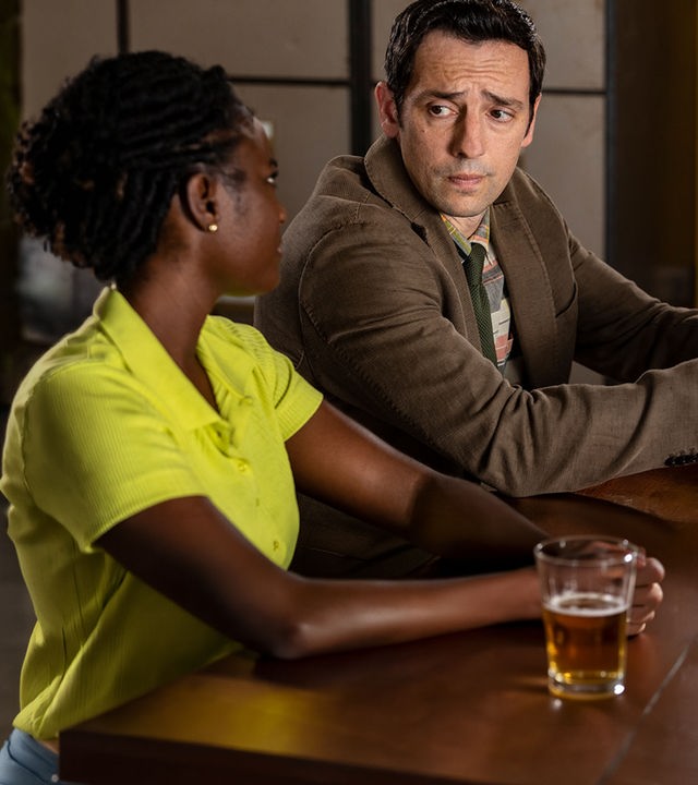 "Death in Paradise - Der Weihnachtsgeist (2)": Naomi Thomas (Shantol Jackson) und Detective Neville Parker (Ralf Little) sitzen nebeneinander an einem Tisch und sehen sich ernst an. Währenddessen hält Detective Parker mit blauen Handschuhen einen beschrifteten Zettel in seiner Hand.