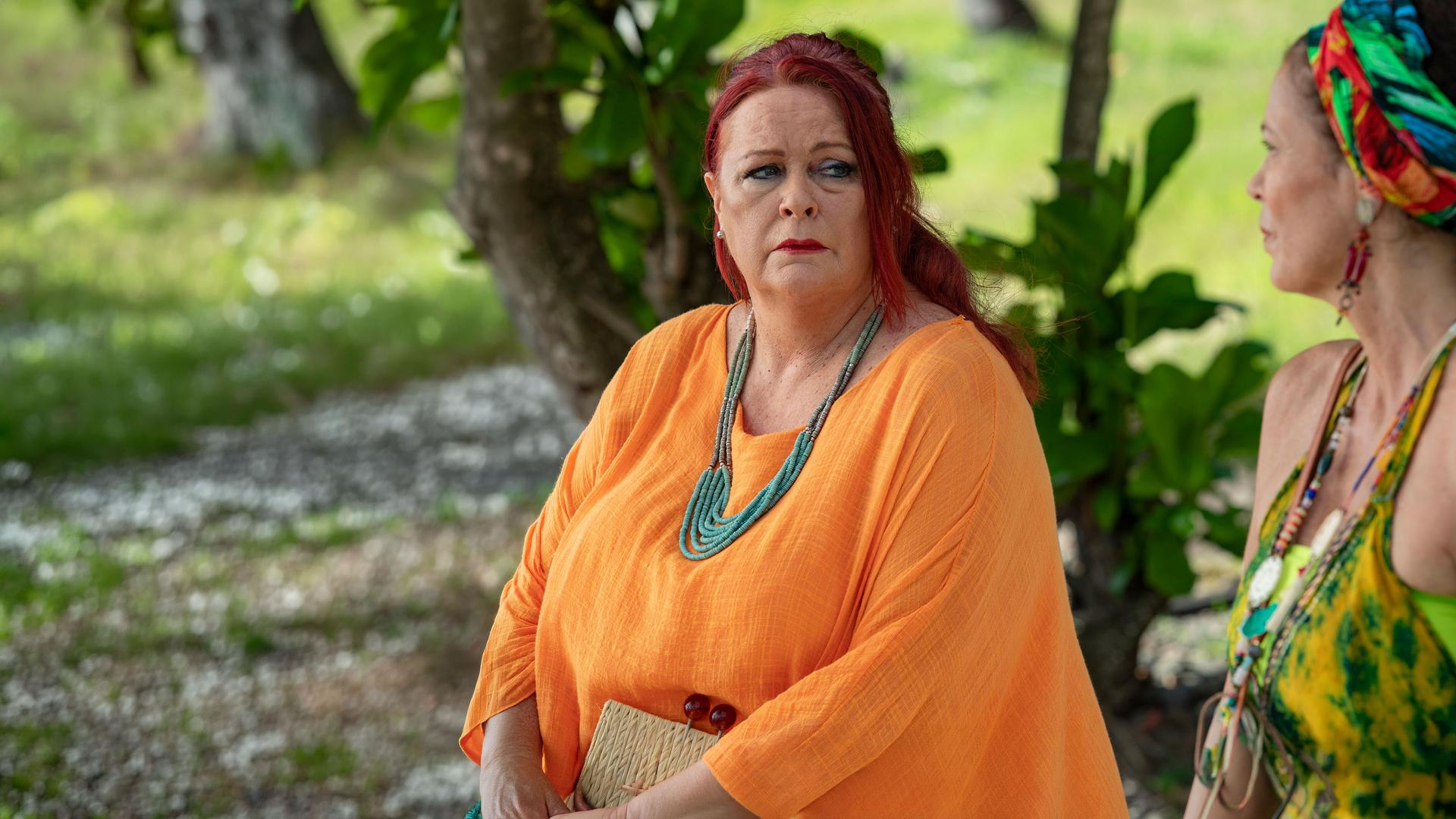 "Death in Paradise - Der Wettbewerb": Catherine Bordey (Elizabeth Bourgine) und Lucky Clayborn (Kate Robbins) sitzen im Grünen nebeneinander. Catherine schaut mit erwartungsvollem Blick zu Lucky, die sie jedoch nur bedrückt ansieht.