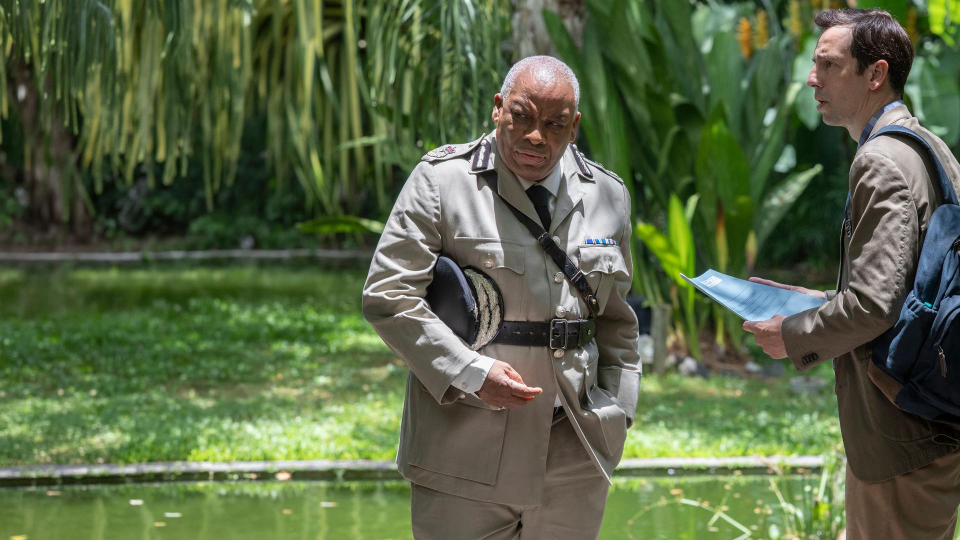 "Death in Paradise - Die letzte Botschaft (2):" Commissioner Selwyn Patterson (Don Warrington) steht im Grünen und blickt mürrisch zur Seite, während DI Neville Parker (Ralf Little) mit einer Polizeiakte in den Händen neben ihm steht und sprachlos in seine Richtung schaut.